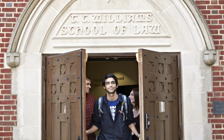 Student with a backpack walking out through the open doors of the T. C. Williams School of Law building, with other students behind him.
