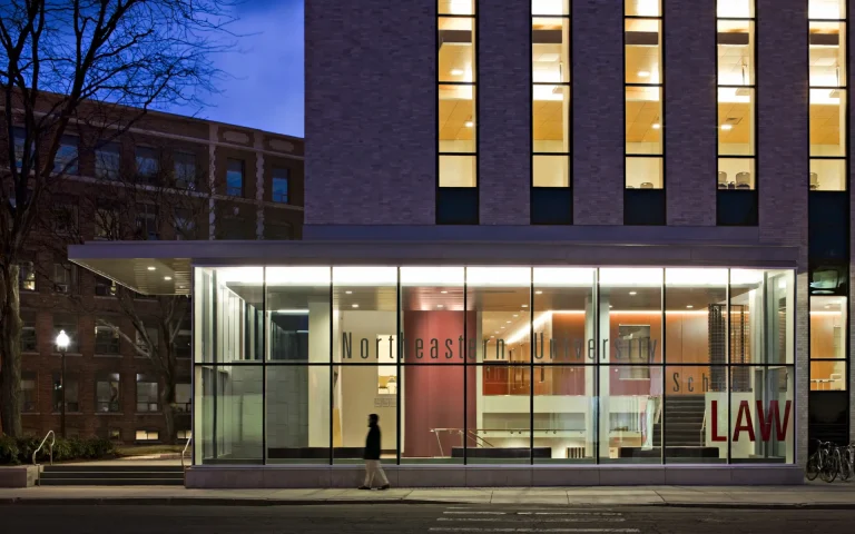 Exterior view of Northeastern University School of Law building at night, with large glass windows showing the lit interior and a person walking along the sidewalk in front.