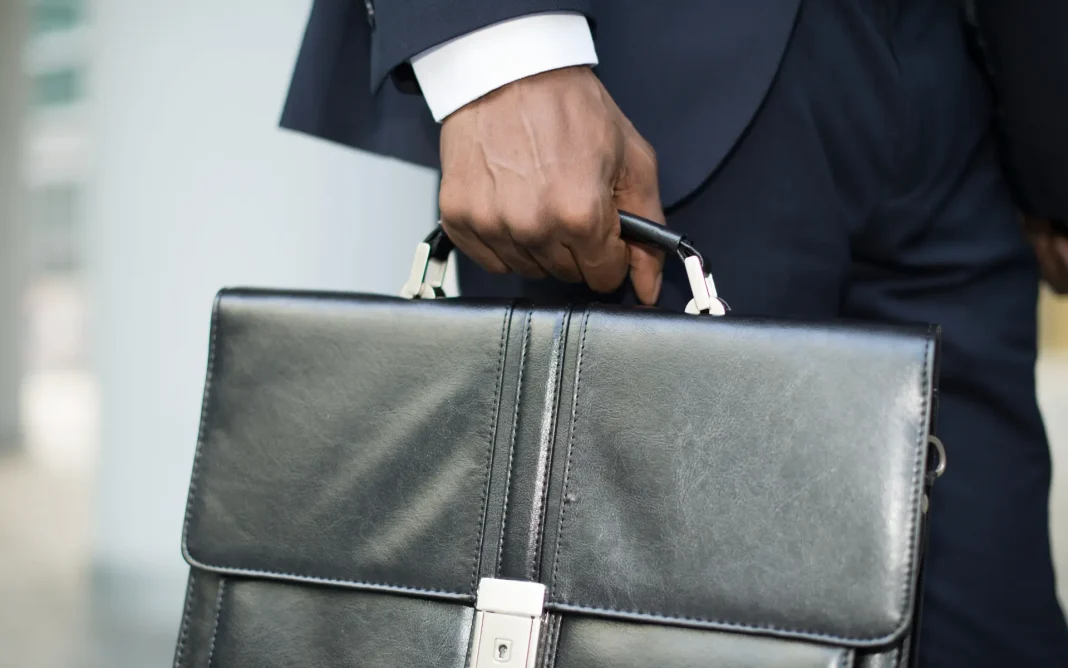 Close-up of a person in a suit holding a black leather briefcase by the handle.