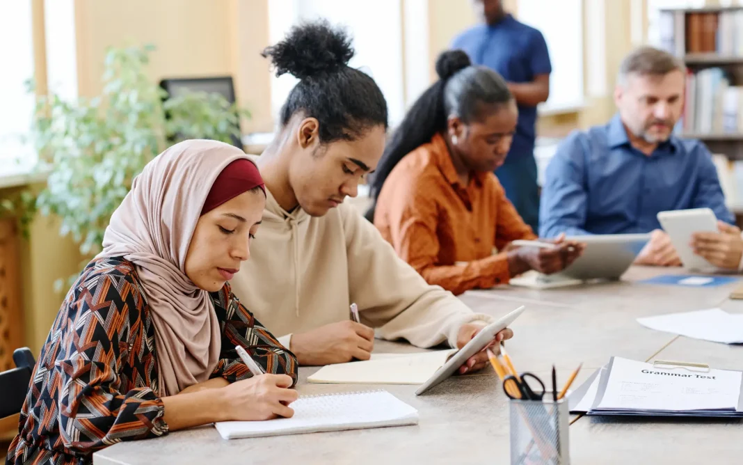 Diverse group of adult students seated at a table in a classroom, writing notes and using tablets during a lesson.