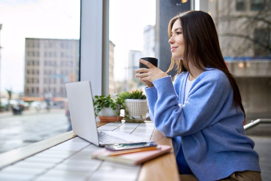 lady holding coffee cup looking out a window with laptop on desk