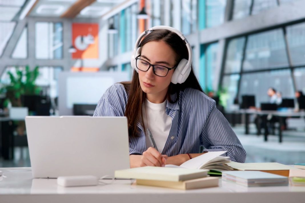 lady with headphones on sitting in library studying in front of a laptop and taking notes