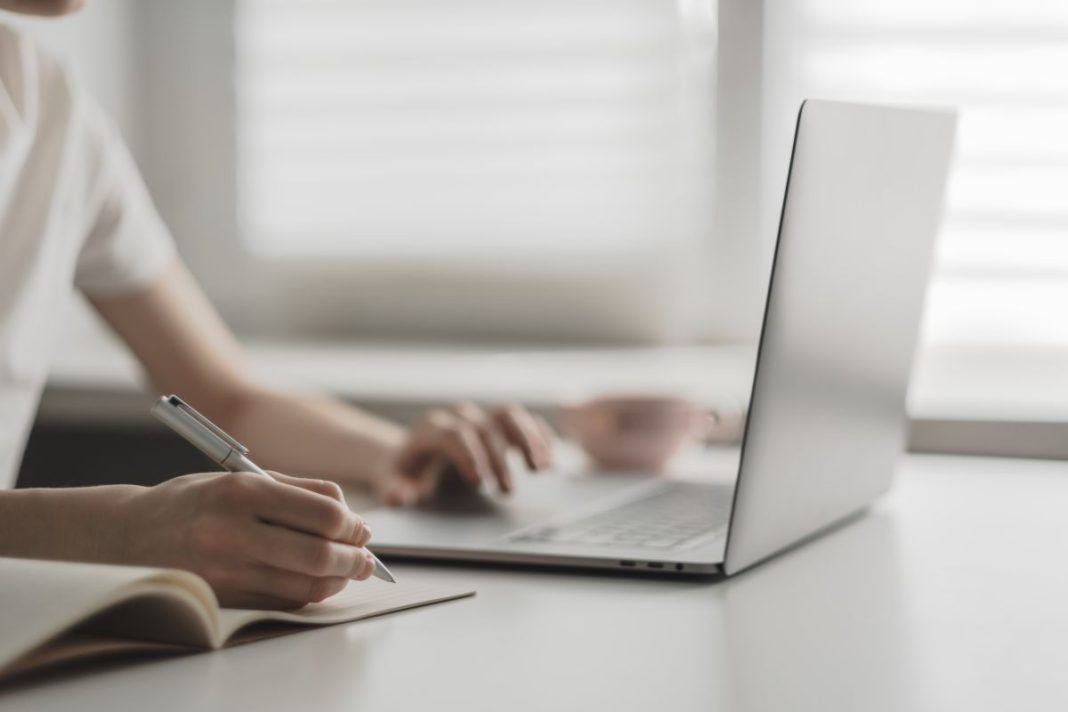 hands of a woman hovering over laptop keyboard and a writing notebook